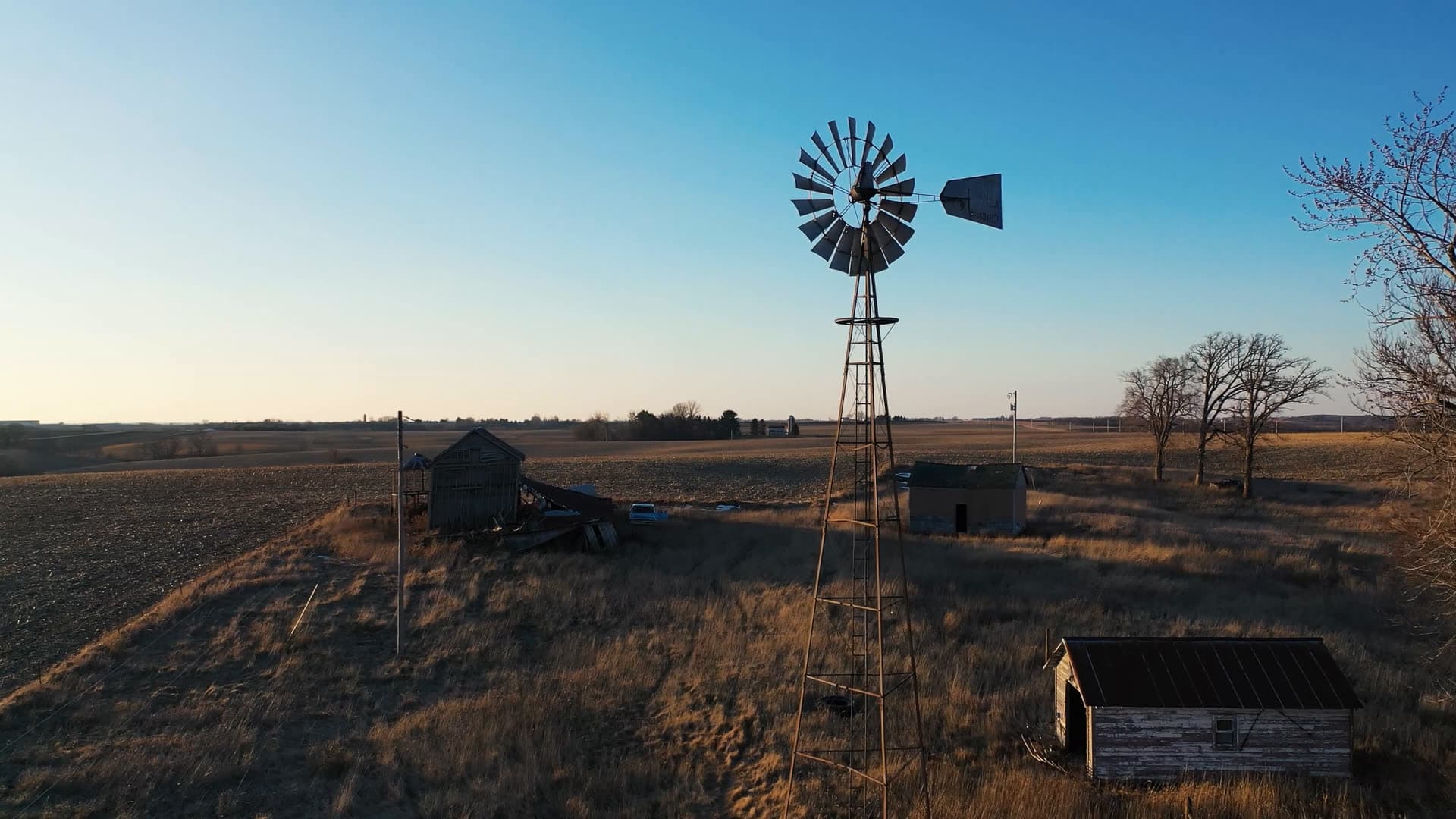 An old farm windmill beside weathered sheds in a dry winter field at sunset