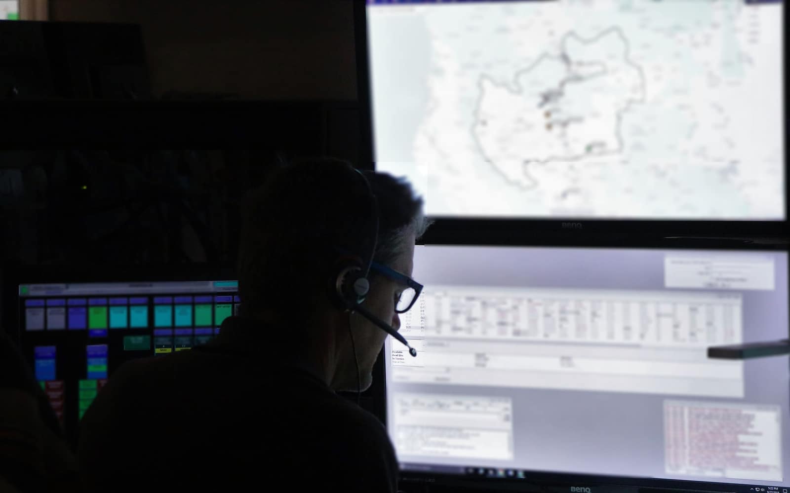 A dispatcher in a headset, seen in silhouette, watches a large map screen in a darkened dispatch center